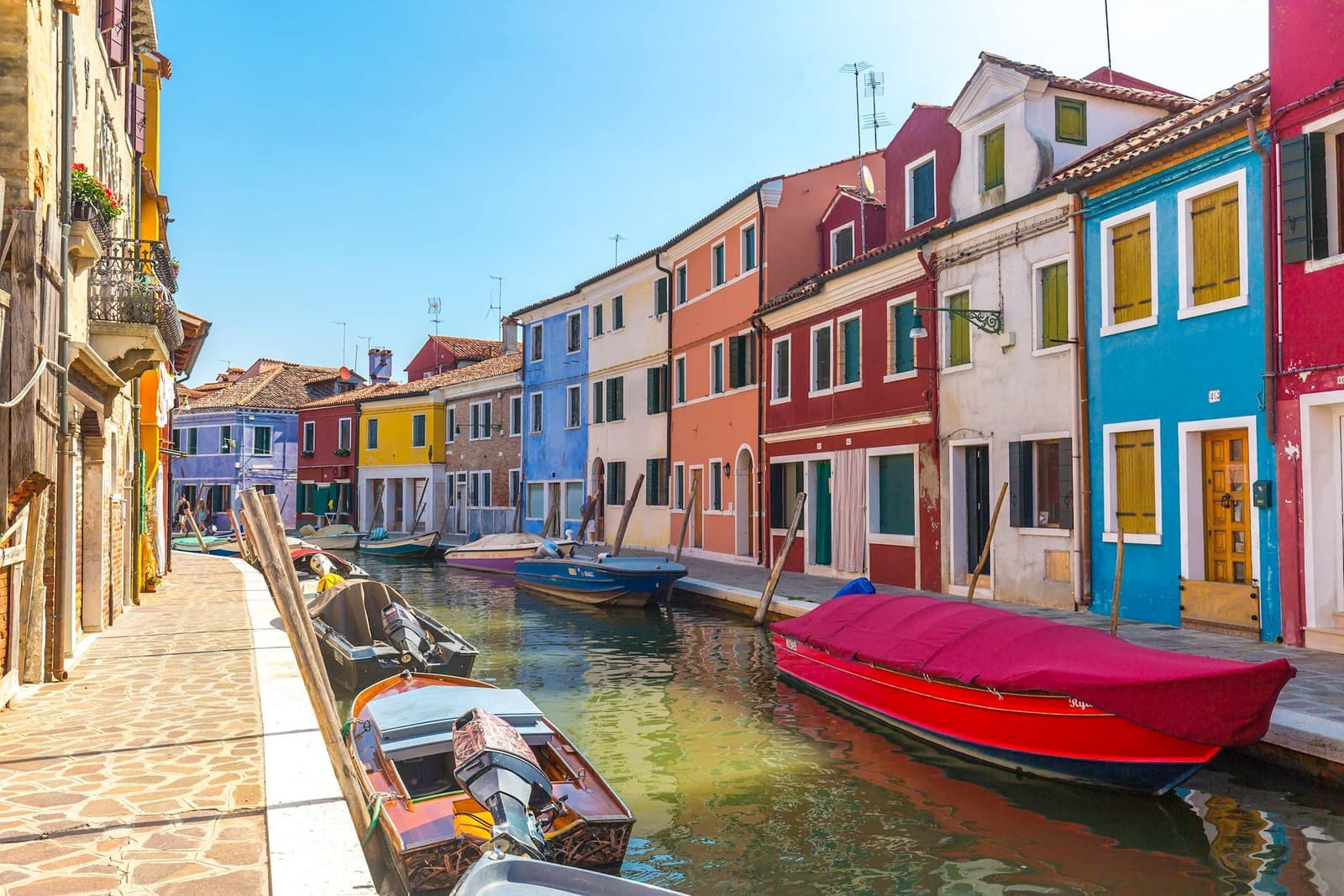 Colourful houses along canal with moored boats and stone walkway in Burano, Venice
