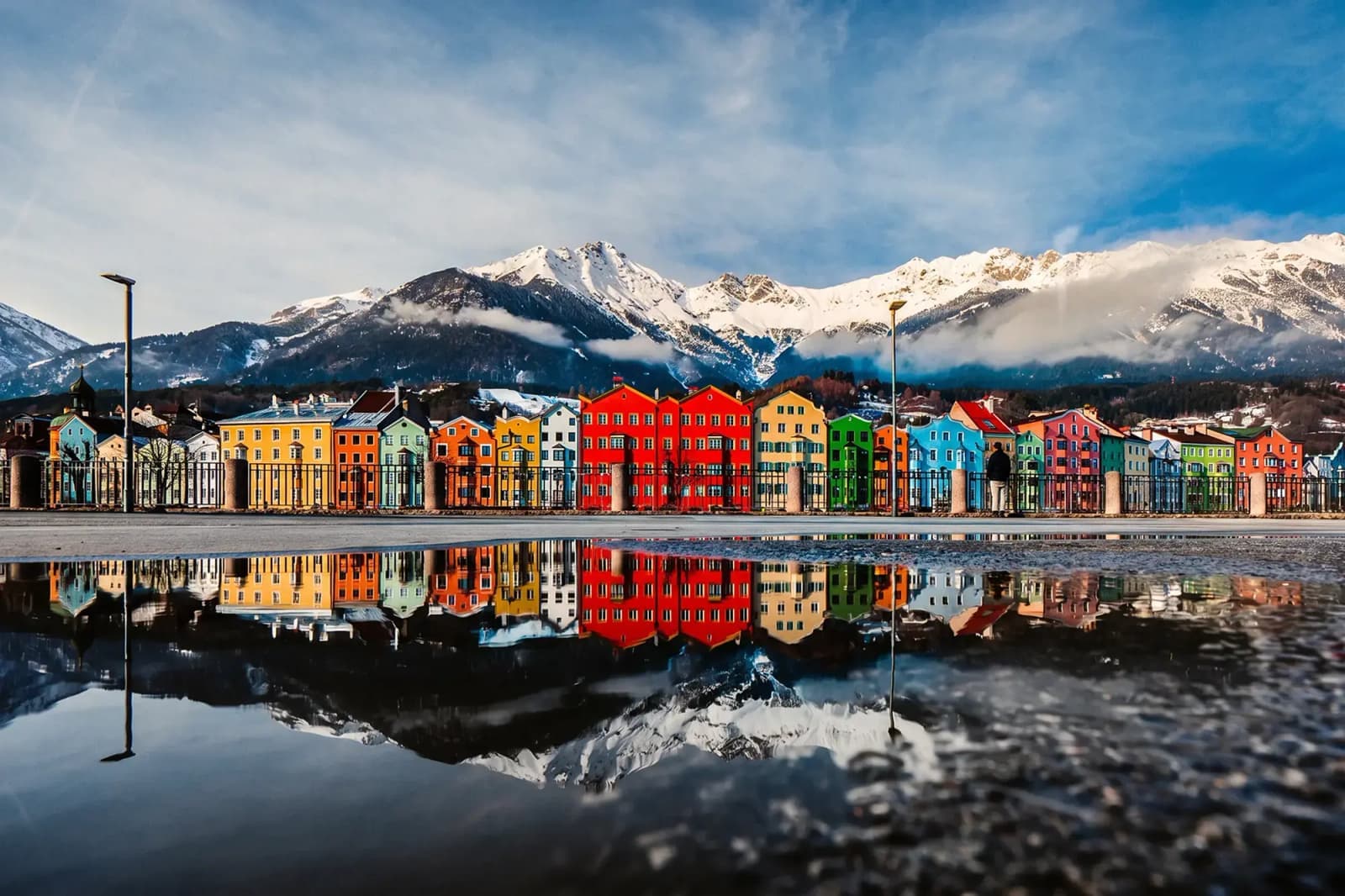 Colorful waterfront buildings reflected in water, with snow-covered mountains in the background.