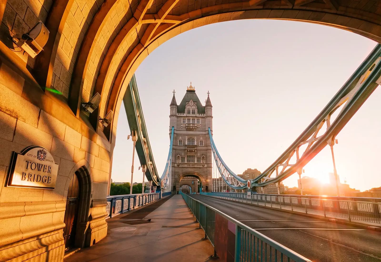A view of London’s Tower Bridge framed by a stone archway on an empty road during sunrise.